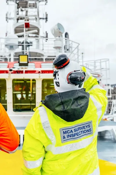 MCA Survey and inspection team member observes a vessel from the deck. They are wearing a yellow hi-visibility jacket and a white hard hat