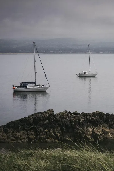 Two small pleasure vessels moored off the coast of Wales. The water is still and the sky is grey and moody